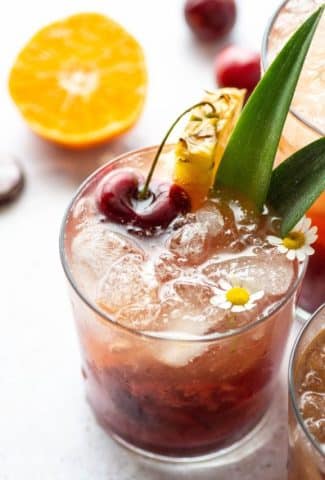 Side view of three tropical kombucha cocktails next to each other with pineapple leaves, pineapple wedges, dark cherries, and sliced tangerine on a light background