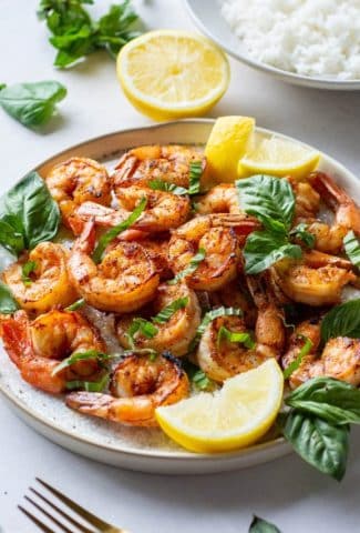 Side angle of a plate of seared shrimp topped with fresh basil and lemon wedges next to a bowl of white rice on a light background