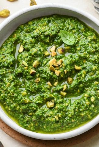 Pistachio pesto in a small shallow ceramic bowl on a cream colored background, surrounded by some scattered pistachios and fresh basil leaves.