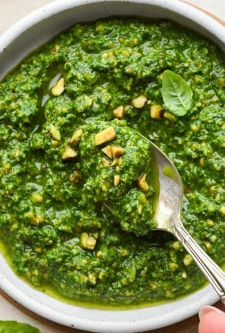 A small bowl of bright green pistachio pesto, topped with a few chopped pistachios and a small basil leaf, with a teaspoon lifting our a small portion of pesto.