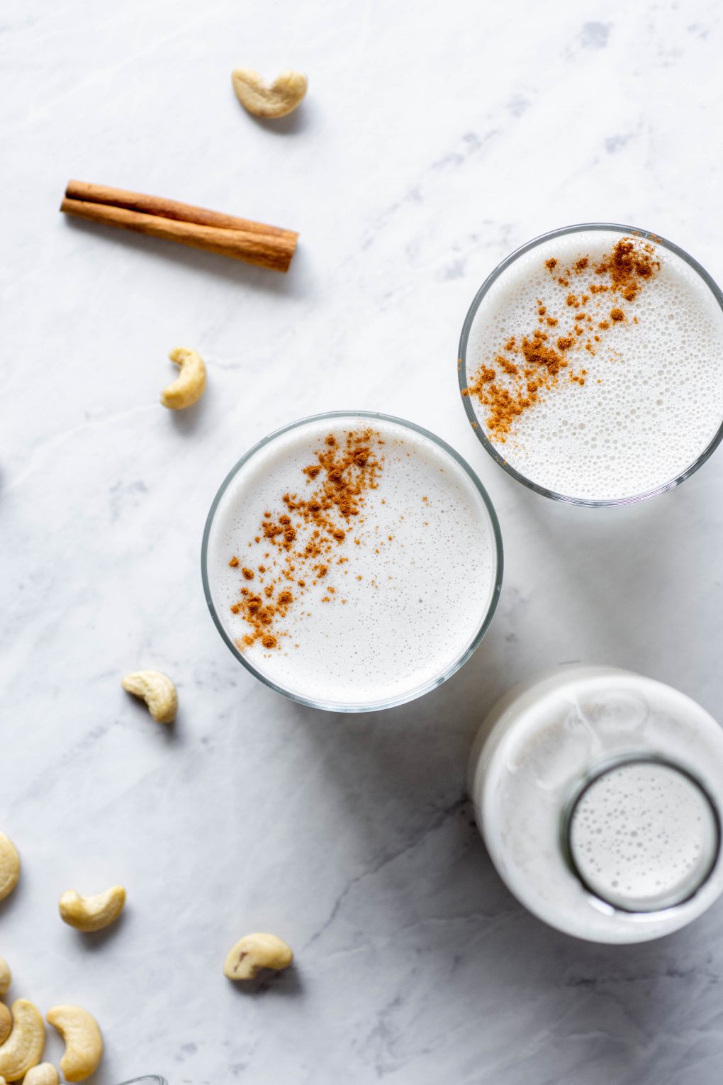 Overhead shot of two side by side glasses of cashew milk sprinkled with cinnamon next to a tall bottle of cashew milk on a marble background next to a cinnamon stick and some scattered cashews