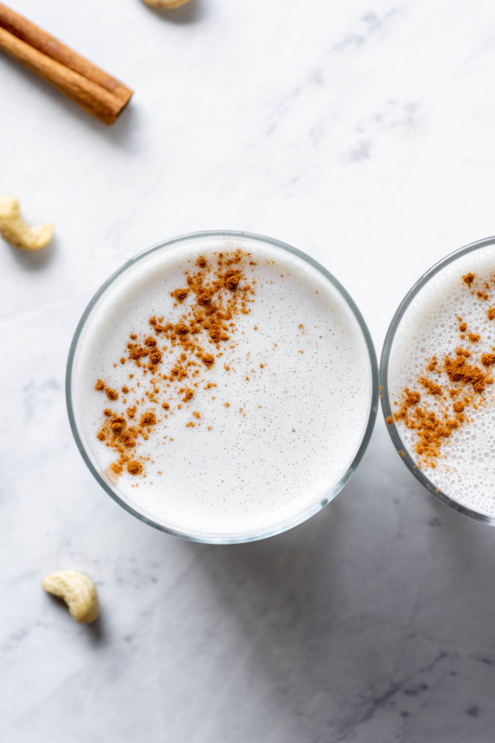 Overhead shot of two side by side glasses of cashew milk sprinkled with cinnamon on a marble background next to a cinnamon stick and some scattered cashews