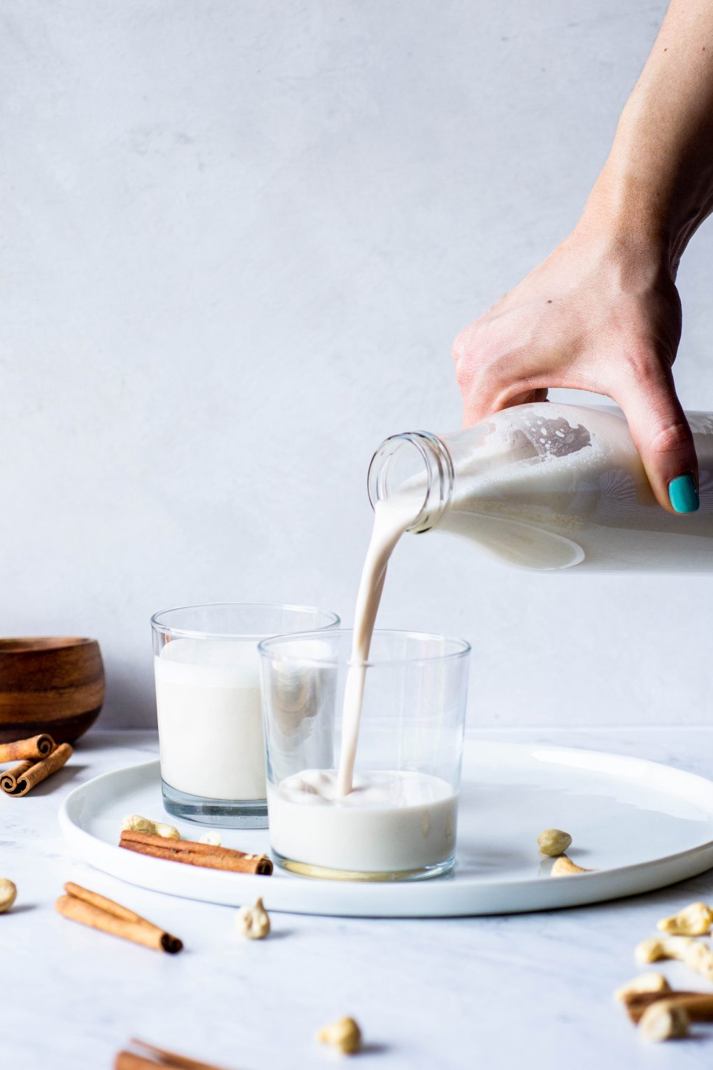 Side view of a hand pouring cashew milk into two glasses on a white plate on a marble background surrounded by cinnamon sticks and cashews