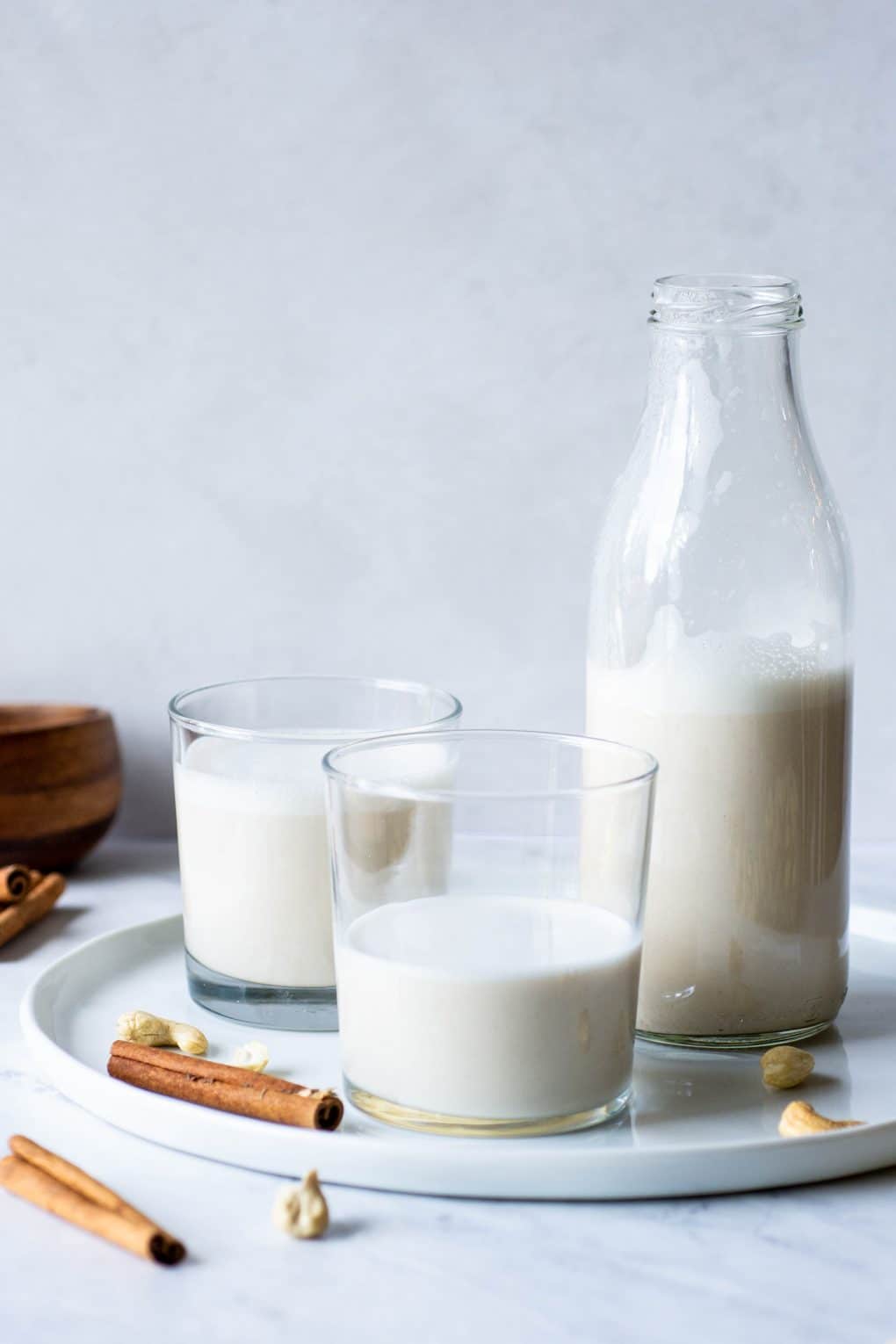 Side view of two cups of cashew milk on a white plate on a marble background surrounded by cinnamon sticks and cashews