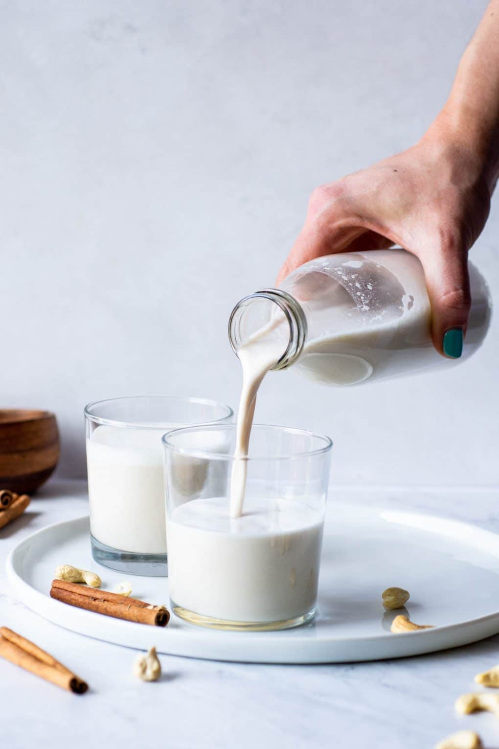 Side view of a hand pouring cashew milk into two glasses on a white plate on a marble background surrounded by cinnamon sticks and cashews