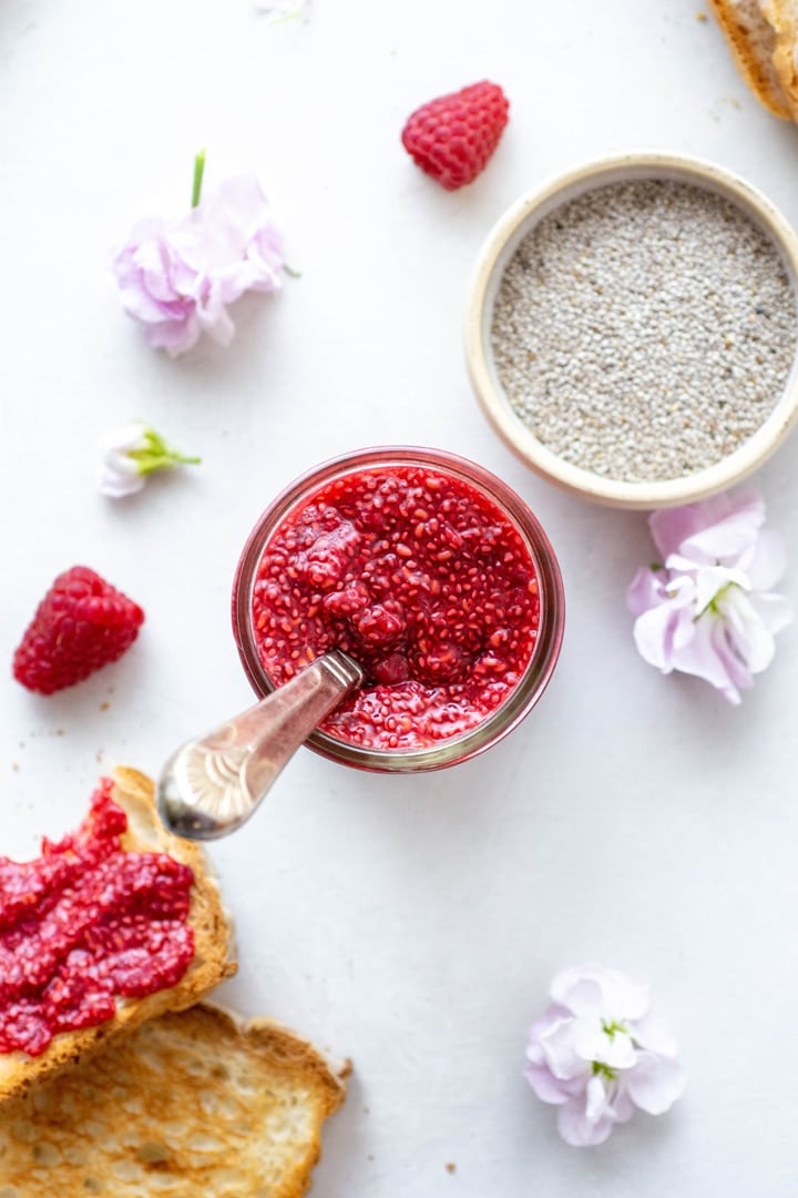 Overhead shot of a jar of raspberry chia seed jam with a spoon inside the jar. On a white background next to a small bowl of white chia seeds, fresh raspberries, and pink flowers.