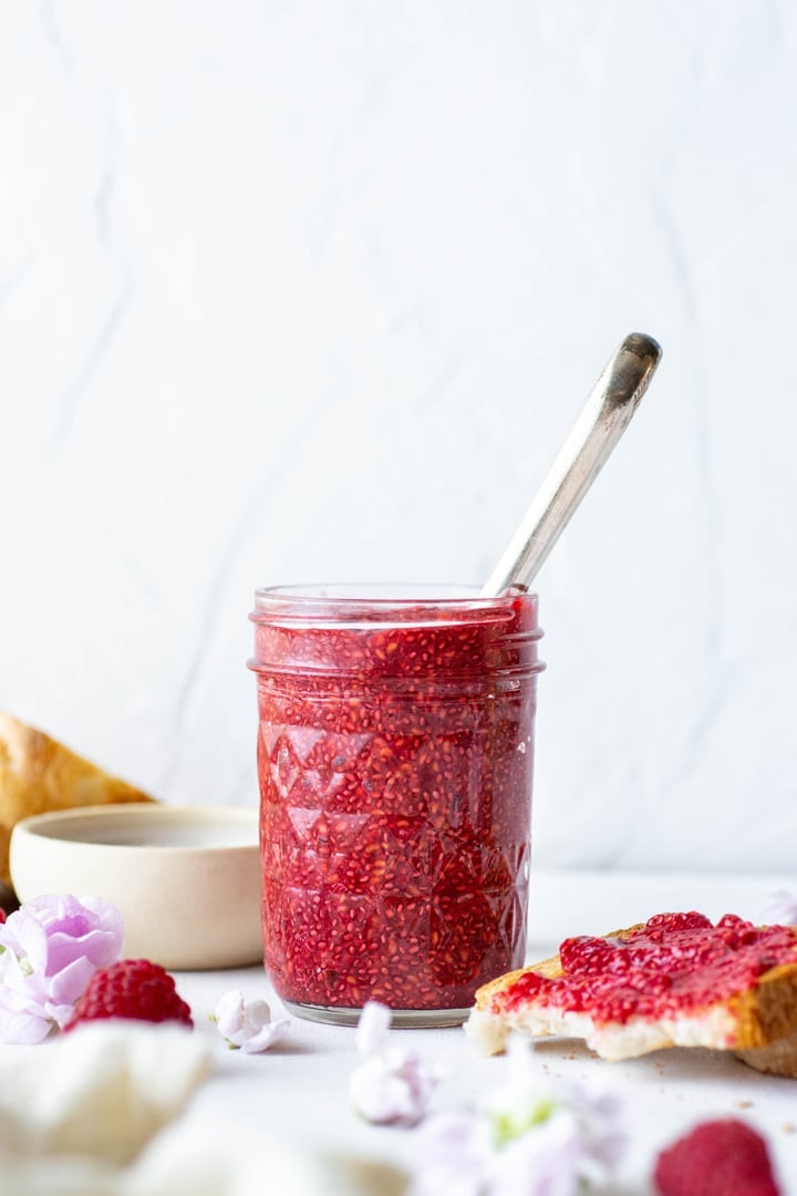 Side view of a jar of raspberry chia seed jam with a spoon inside the jar. On a white background next to a small bowl of white chia seeds, fresh raspberries, toast spread with jam, and pink flowers.