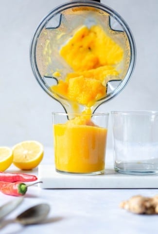 Pouring a bright yellow turmeric slushy into a short clear glass against a white background