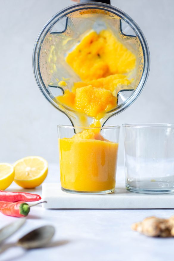 Pouring a bright yellow turmeric slushy into a short clear glass against a white background