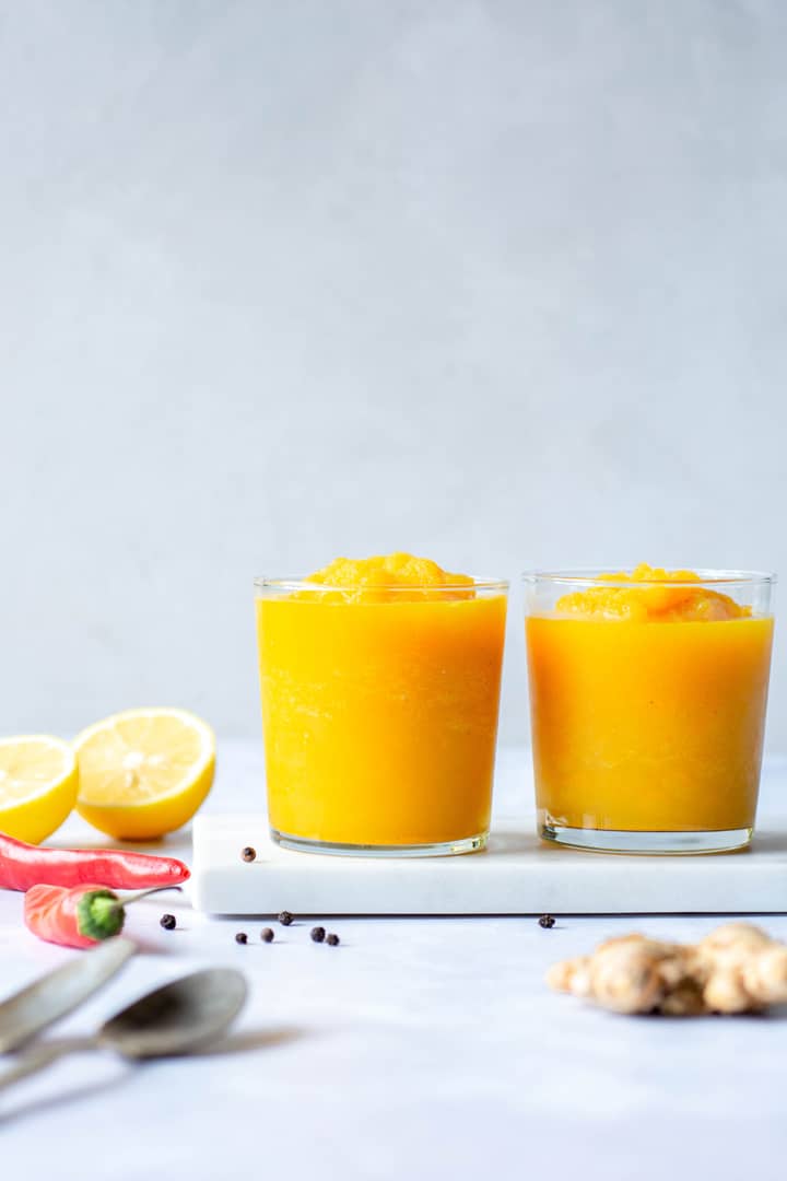 Two side by side short clear glasses of a bright yellow turmeric slushy against a white background surrounded be lemon halves, chili peppers, ginger, and black peppercorns