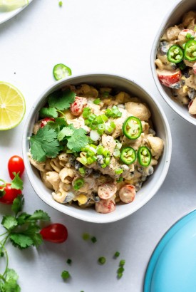 A close up shot of a bowl of vegan mexican mac and cheese topped with green onions, jalapeno, and fresh cilantro. On a light background surrounded by sliced lime, cherry tomatoes, and fresh herb.