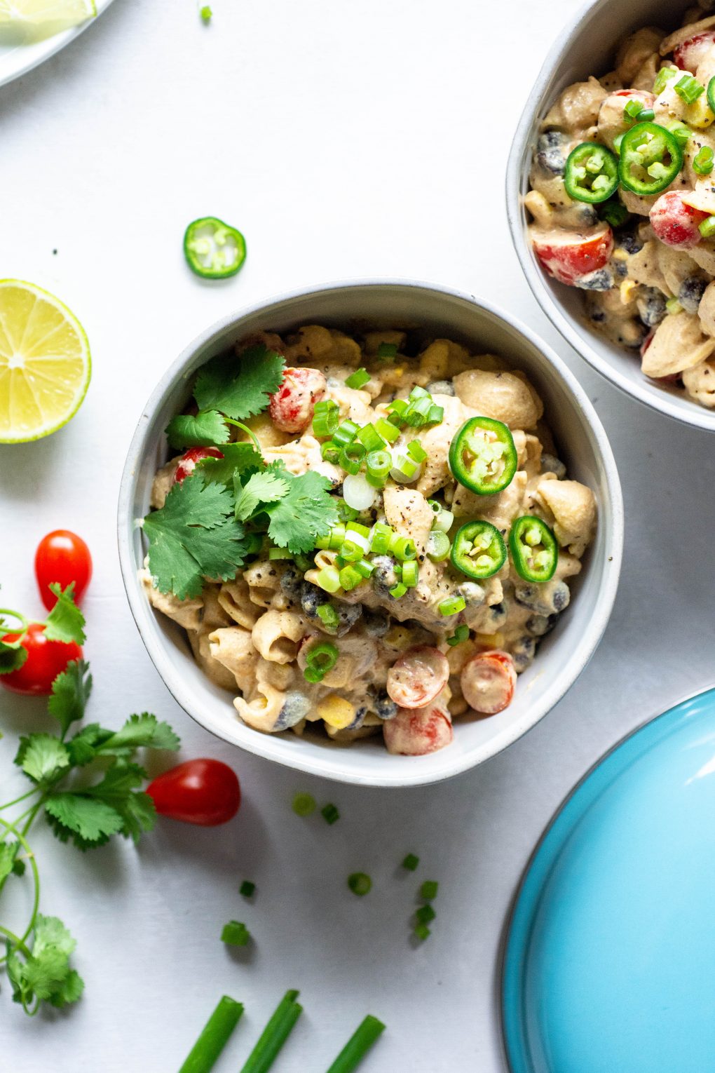 Two side by side bowls of vegan mexican mac and cheese topped with green onions, jalapeno, and fresh cilantro. On a light background surrounded by sliced lime, cherry tomatoes, and fresh herb.