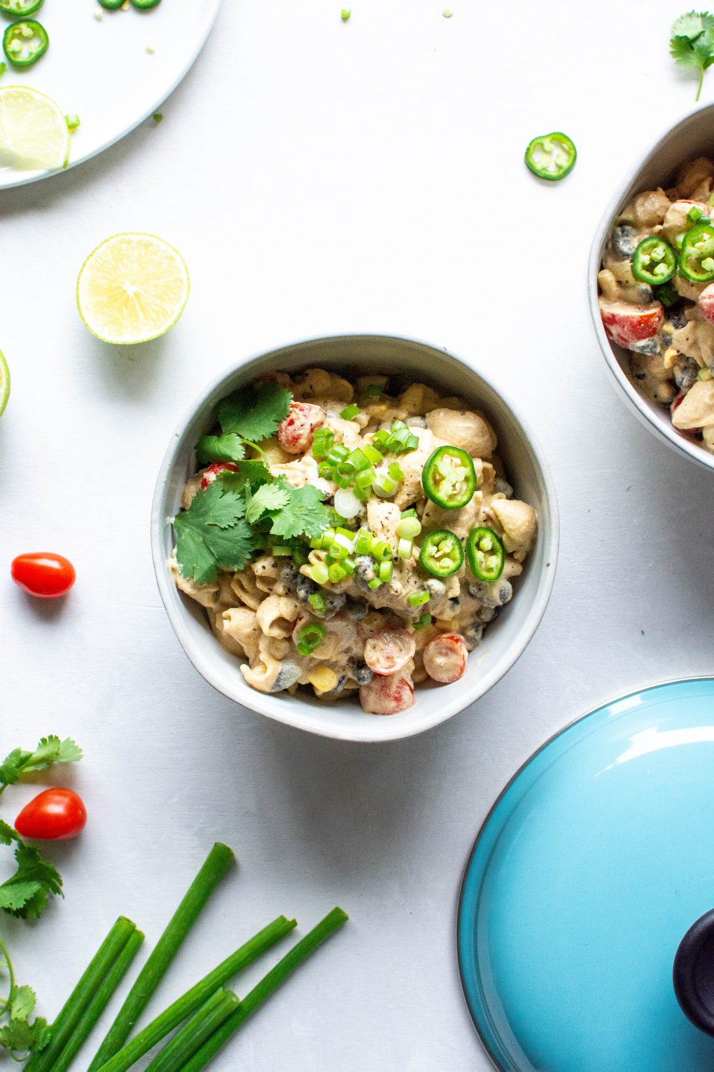 Two side by side bowls of vegan mexican mac and cheese topped with green onions, jalapeno, and fresh cilantro. On a light background surrounded by sliced lime, cherry tomatoes, and fresh herb.