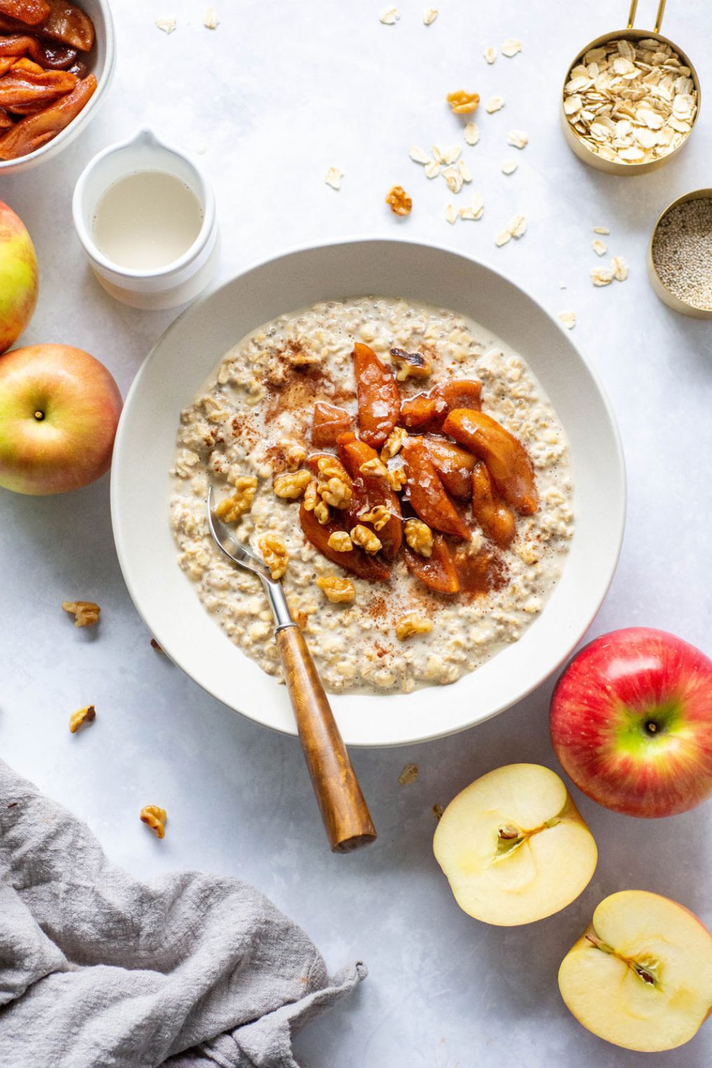 Large white bowl of salted caramel apple overnight oats topped with a big scoop of baked cinnamon apples, walnuts, and flaky sea salt. Bowl is on a light background surounded by cut apples, and bits of walnuts.