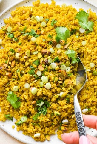 Curried quinoa pilaf on a large serving platter, topped with cilantro, pistachios, and green onions, with a serving spoon lifting some off the plate.