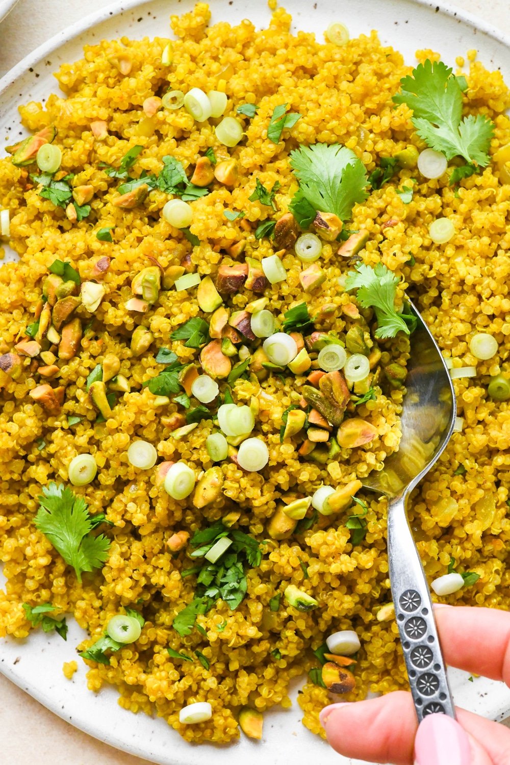 Curried quinoa pilaf on a large serving platter, topped with cilantro, pistachios, and green onions, with a serving spoon lifting some off the plate.