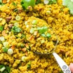 Curried quinoa pilaf on a large serving platter, topped with cilantro, pistachios, and green onions, with a close up of a serving spoon lifting some quinoa off the plate.