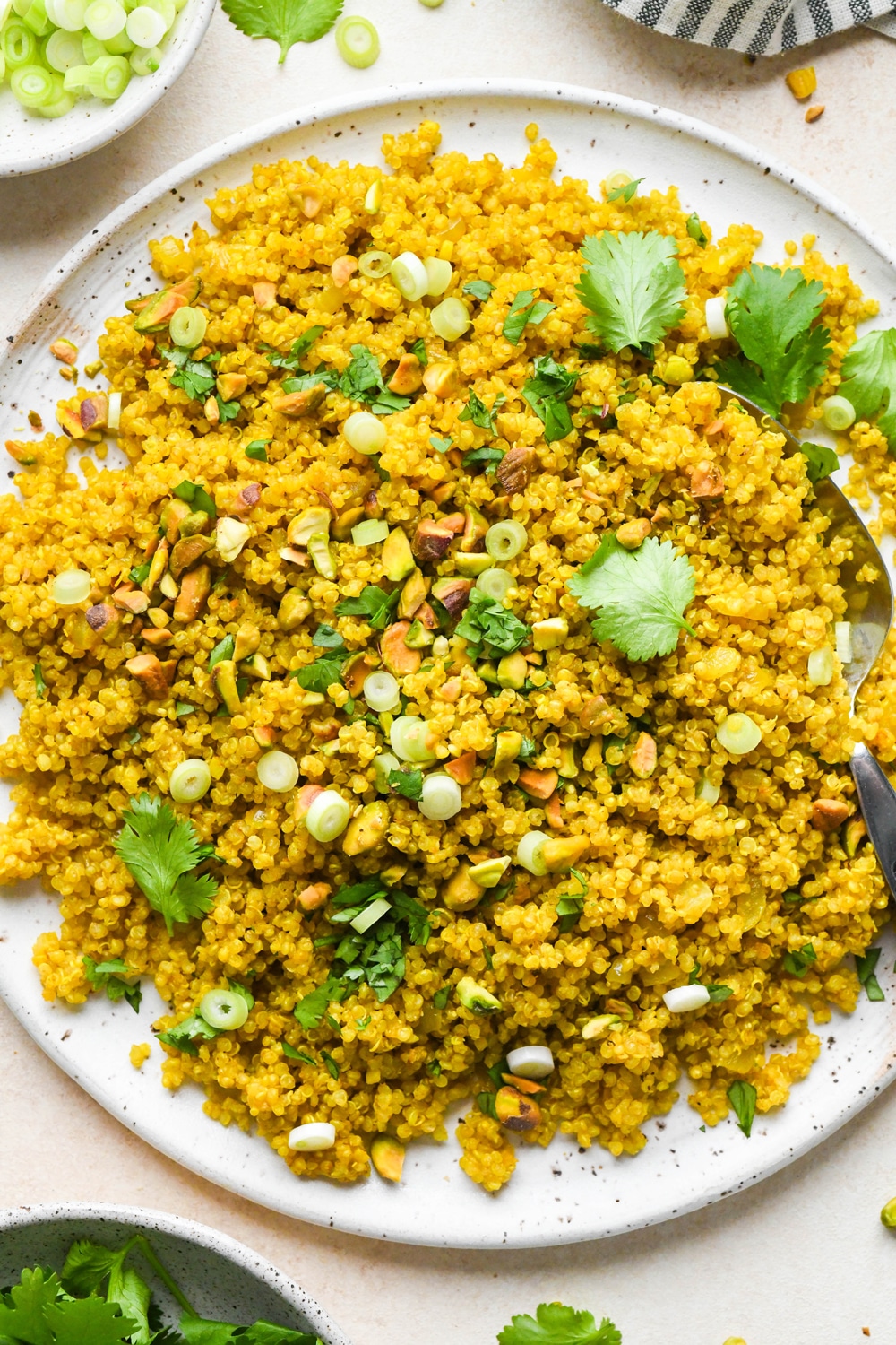 Curried quinoa pilaf on a large serving platter topped with chopped pistachios, fresh cilantro, and thinly sliced green onion next to a striped linen napkin, a small dish of chopped pistachios, and a small dish of fresh cilantro.