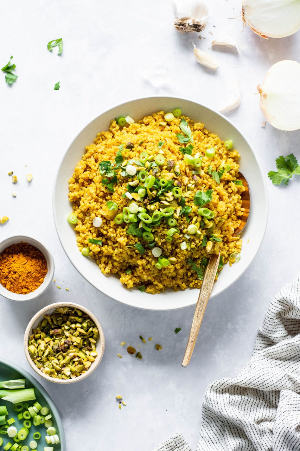 Big white bowl of bright yellow curried quinoa topped with cilantro, green onions, and pistachios. On a light colored background next to a small bowl of pistachios, a small bowl of curry powder, and an onion sliced in half. There's a large gold metal spoon in the bowl. 
