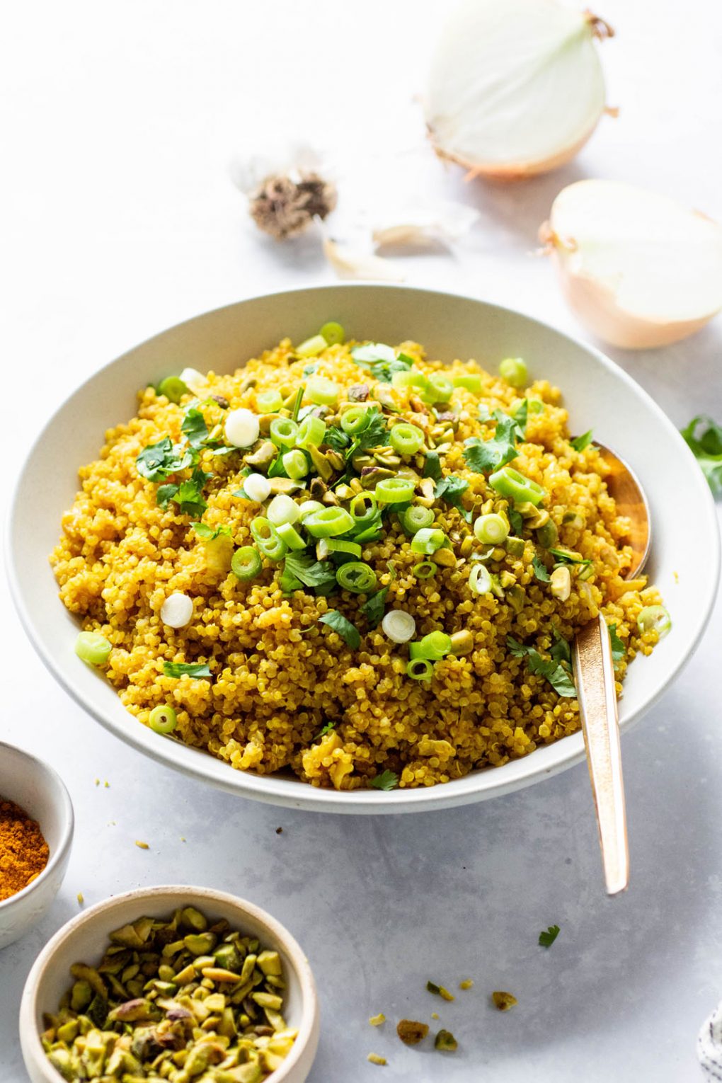 Side angle view of a big white bowl of bright yellow curried quinoa topped with cilantro, green onions, and pistachios. On a light colored background next to a small bowl of pistachios.