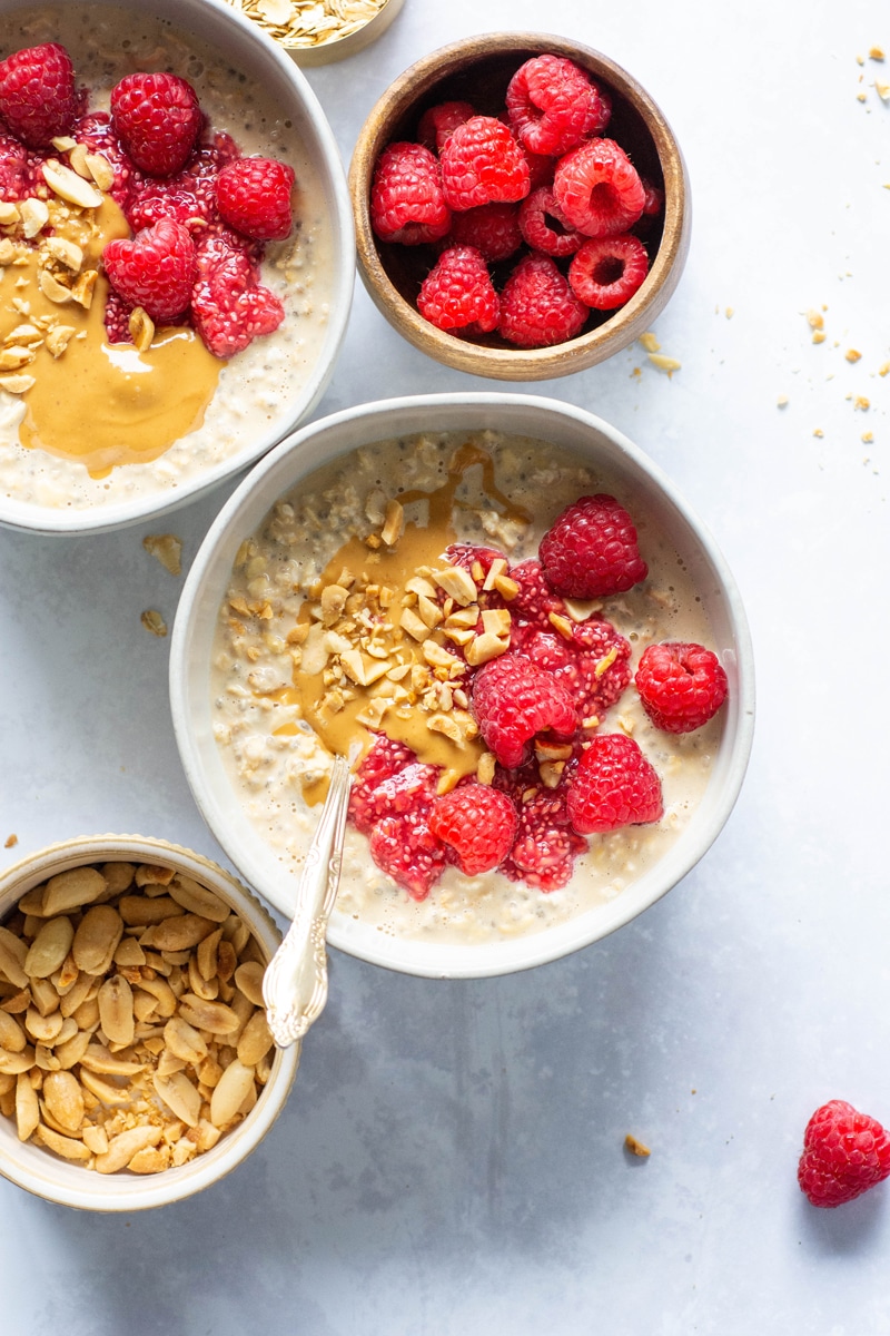 2 small white bowls of peanut butter overnight oats topped with a peanut butter drizzle, crushed peanuts, raspberry chia seed jam and fresh raspberries. In between a smaller bowl of raspberries, and a smaller bowl of peanuts. 