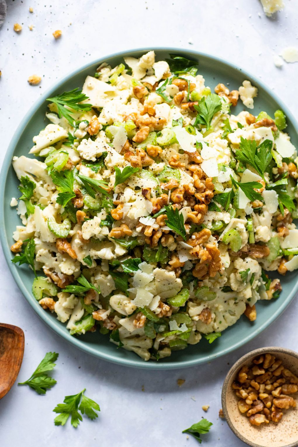 A large round plate with shaved cauliflower and celery salad topped with walnuts, parsley, and shave cheese. On a light background surrounded by bits of nuts, cheese, and parsley. Wooden salad serving utensils lie next to the plate. 