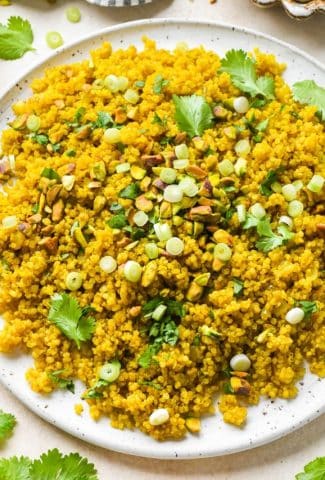 Curried quinoa pilaf on a large serving platter topped with chopped pistachios, fresh cilantro, and thinly sliced green onion next to a striped linen napkin and a small dish of chopped pistachios.