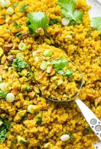 Curried quinoa pilaf on a large serving platter, topped with cilantro, pistachios, and green onions, with a close up of a serving spoon lifting some quinoa off the plate.
