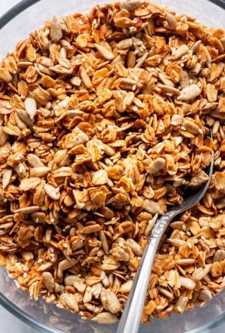 Close up overhead shot of a large glass pyrex dish full of toasty granola with a silver spoon angled in. On a white background.
