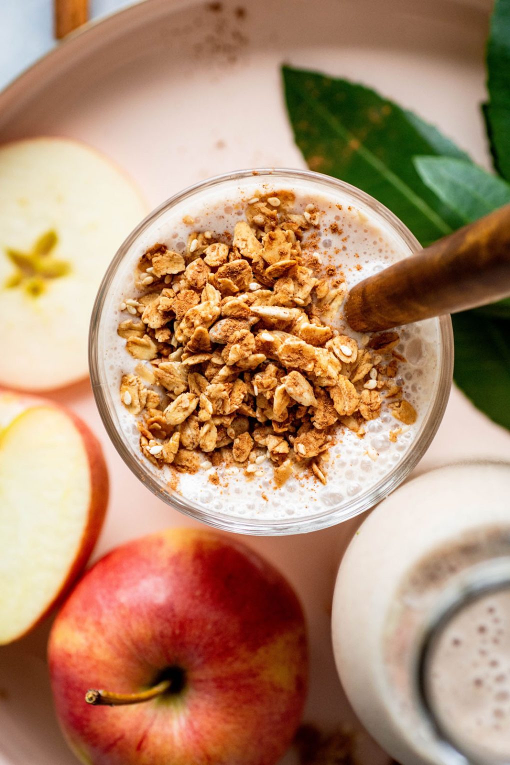 Close up over head shot of a glass full of cinnamon apple pie smoothie with a wooden handled spoon. Sitting in a gold rimmed round serving tray surrounded by apples, scattered granola, greenery, and cinnamon sticks.