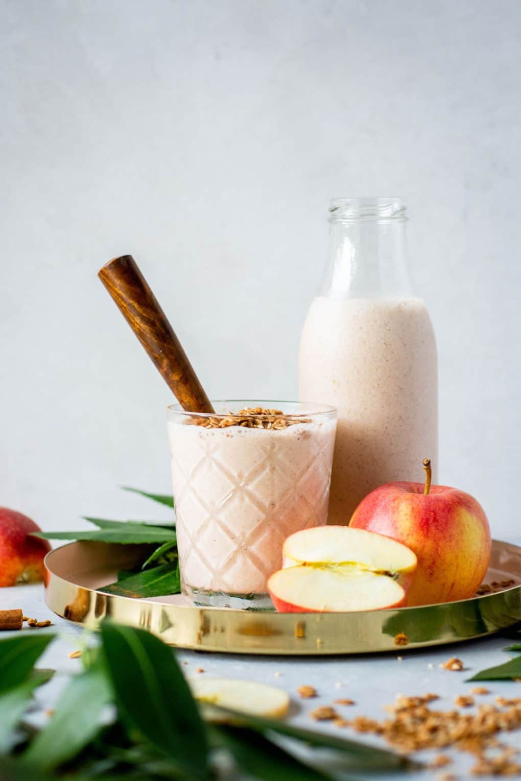 Glass full of cinnamon apple pie smoothie with a wooden handled spoon. Sitting in a gold rimmed round serving tray next to a larger jar of smoothie and surrounded by apples, scattered granola, greenery, and cinnamon sticks.