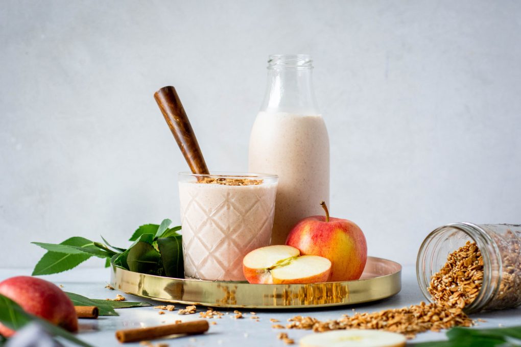 Glass full of cinnamon apple pie smoothie with a wooden handled spoon. Sitting in a gold rimmed round serving tray next to a larger jar of smoothie and surrounded by apples, an over tipped jar of granola, greenery, and cinnamon sticks.