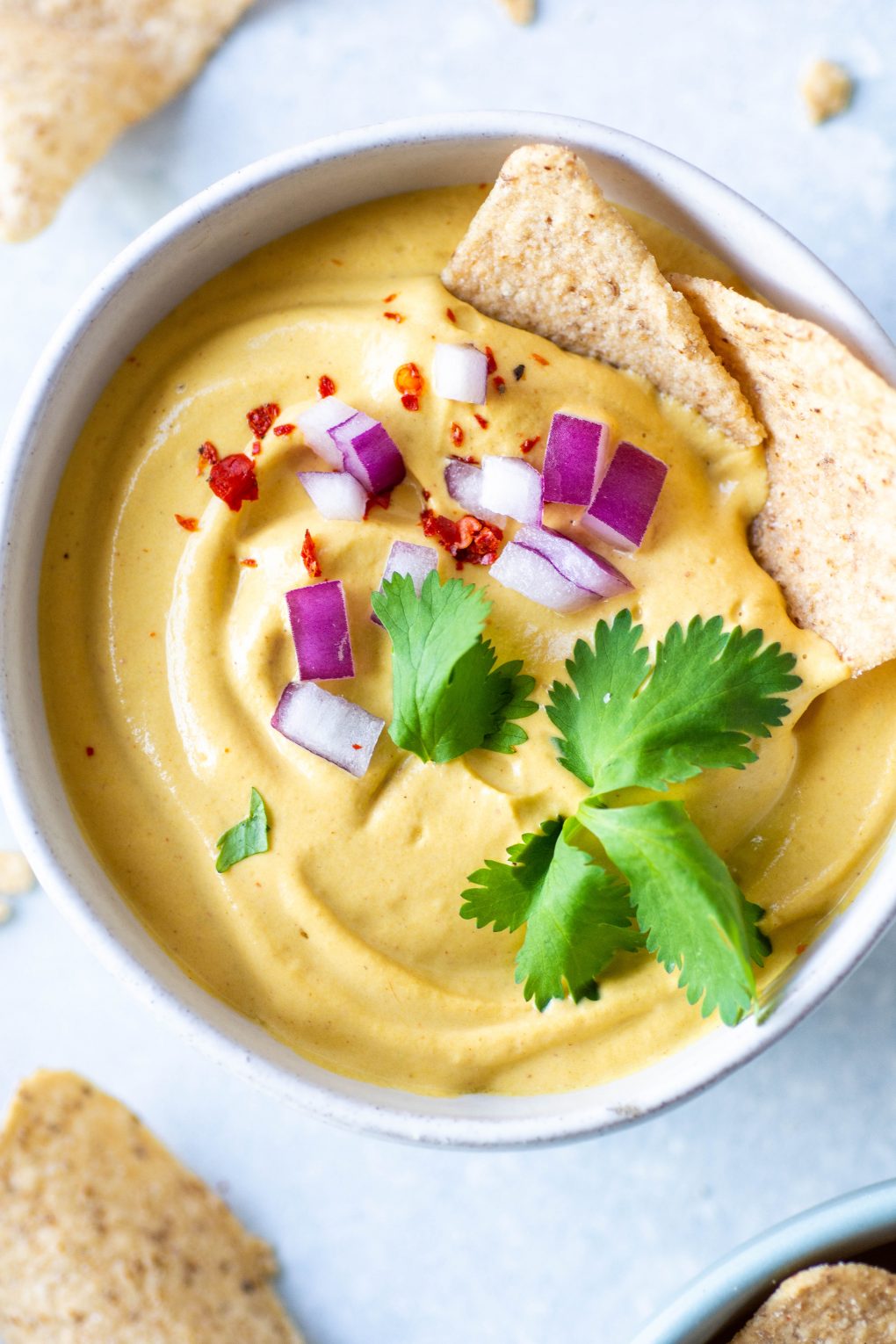 Close up shot of a small white bowl of pumpkin cashew queso on a light background. Garnished with chopped red onion, cilantro, and red chili flakes. Two chips are half dipped into the queso and it's surrounded by lime wedges and scattered chips.