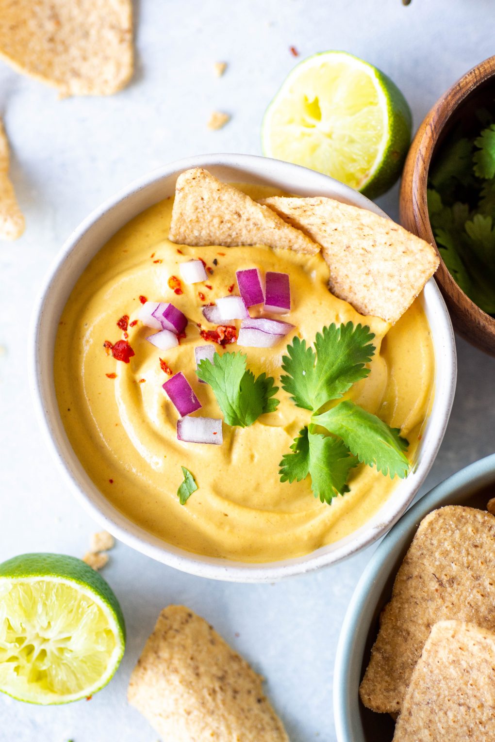 Small white bowl of pumpkin cashew queso on a light background. Garnished with chopped red onion, cilantro, and red chili flakes. Two chips are half dipped into the queso and it's surrounded by lime wedges and scattered chips.
