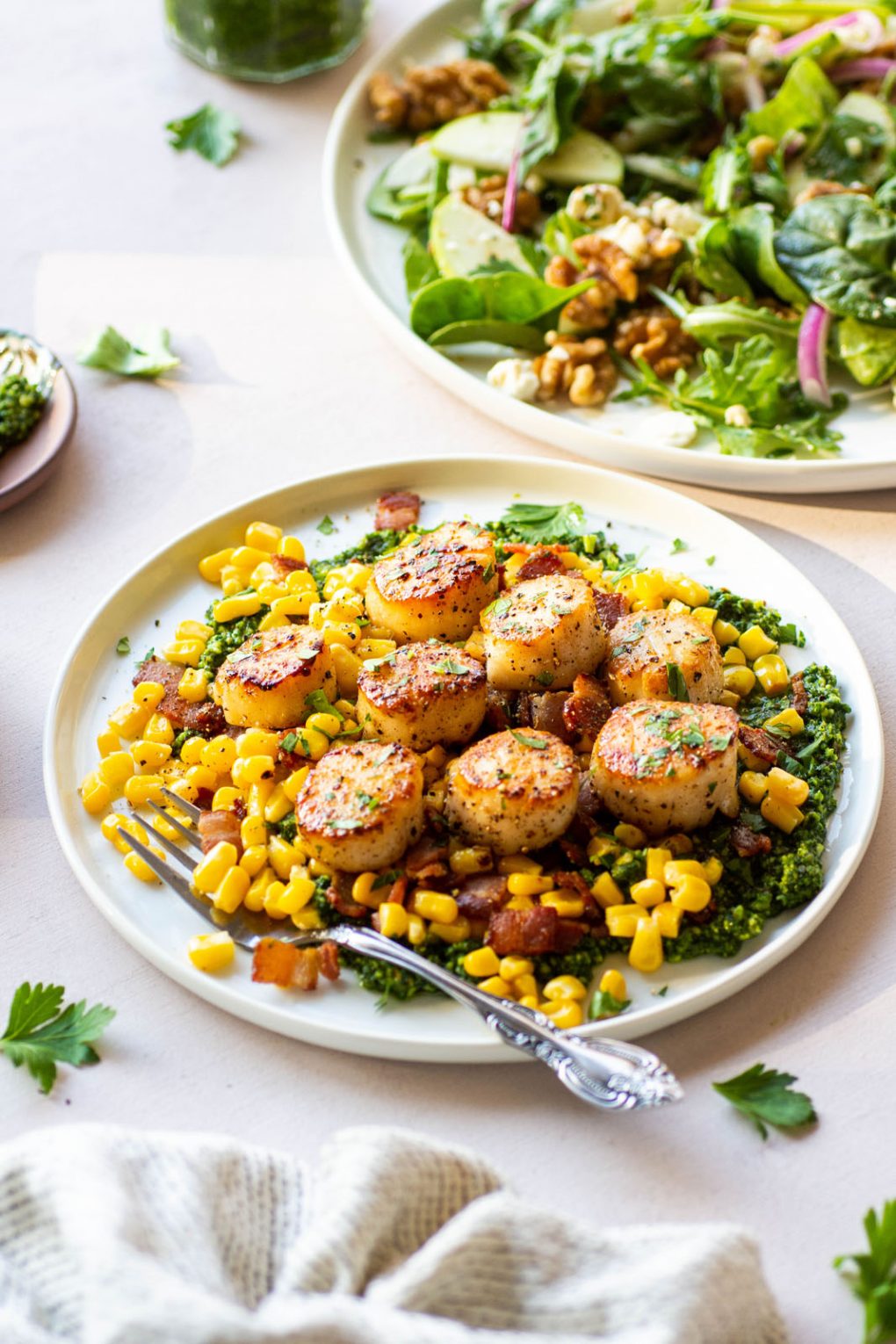 Zoomed out side angle shot of scallops over a corn and bacon saute and kale pesto. On a round white plate placed on a light colored background. In the background is a large plate of salad and some torn parsley leaves.