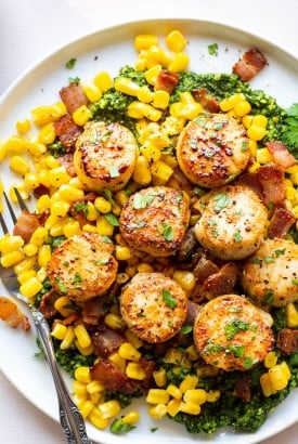 Perfectly seared scallops over a corn and bacon saute and kale pesto. On a round white plate placed on a light colored background, with a fork on the edge of the plate.