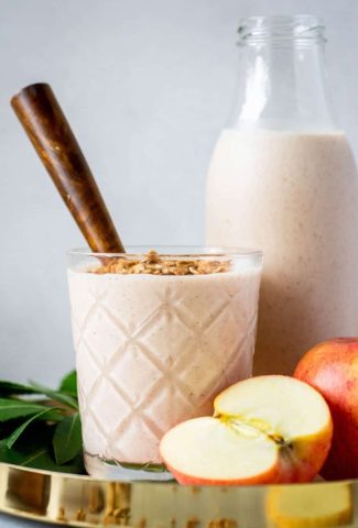 Close up shot of a glass full of cinnamon apple pie smoothie with a wooden handled spoon. Sitting in a gold rimmed round serving tray next to a larger jar of smoothie and surrounded by apples, scattered granola, greenery, and cinnamon sticks.