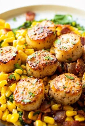 Close up side angle shot of perfectly seared scallops over a corn and bacon saute and kale pesto. On a round white plate placed on a light colored background.