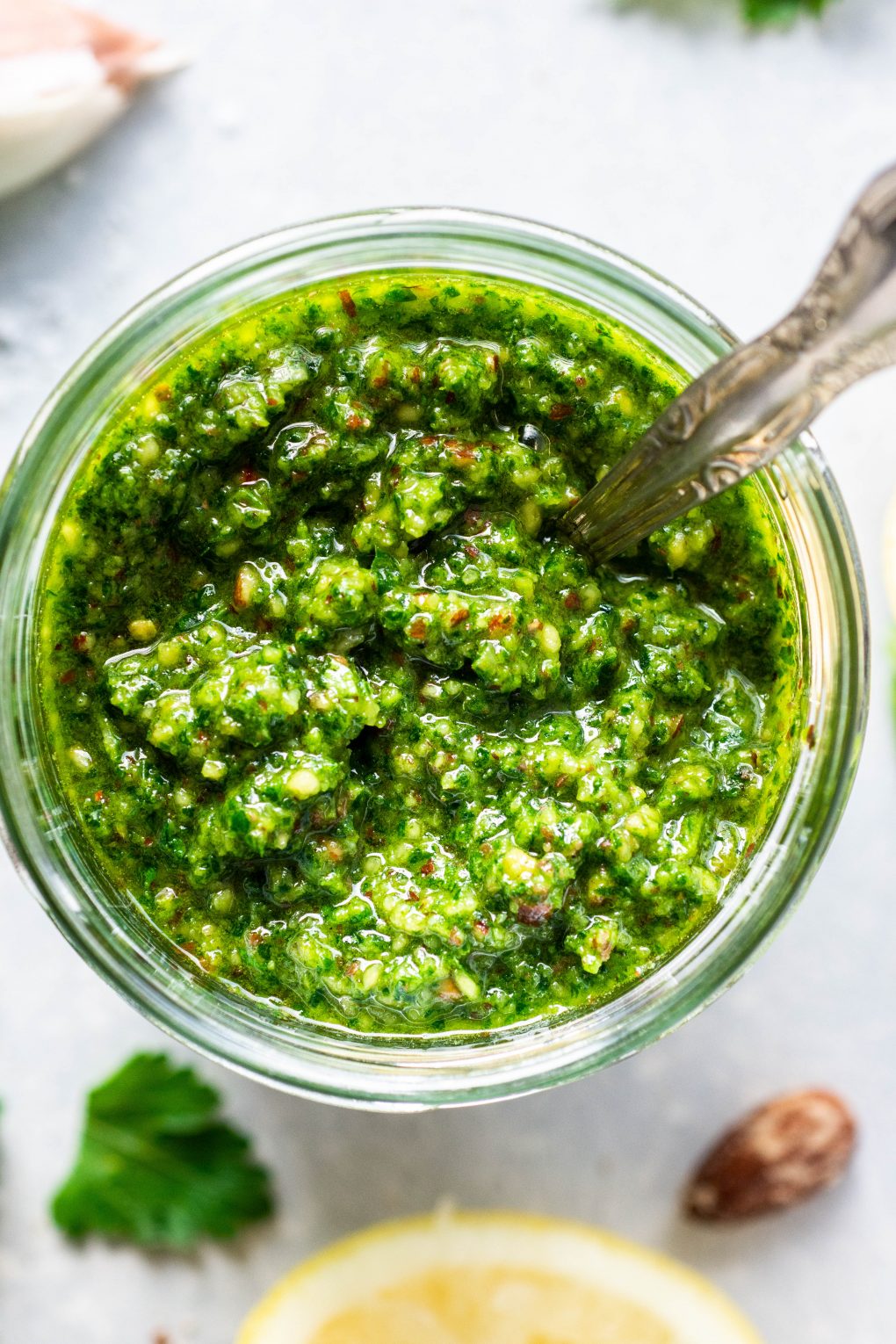 Close up shot of a glass jar of bright green kale pesto with a spoon. On a light colored background next to some fresh parsley leaves.