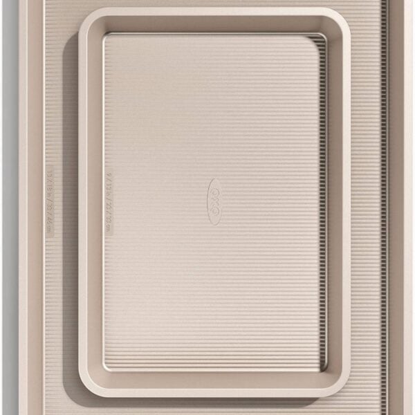 Two beige, non-stick baking sheets of two sizes are stacked inside each other on a white background. The pans have slightly raised edges and a textured surface.