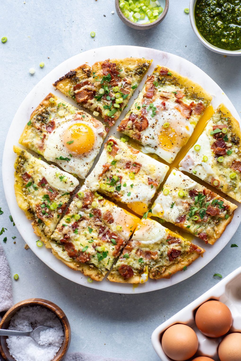 Overhead shot of a breakfast pizza cut into 9 squares. Topped with melted cheese, crispy bacon, sunny side up eggs, and fresh herbs and green onions. On a light colored marble serving tray and a light background. Next to a small bowl of sliced green onions, and another small bowl of pesto. 