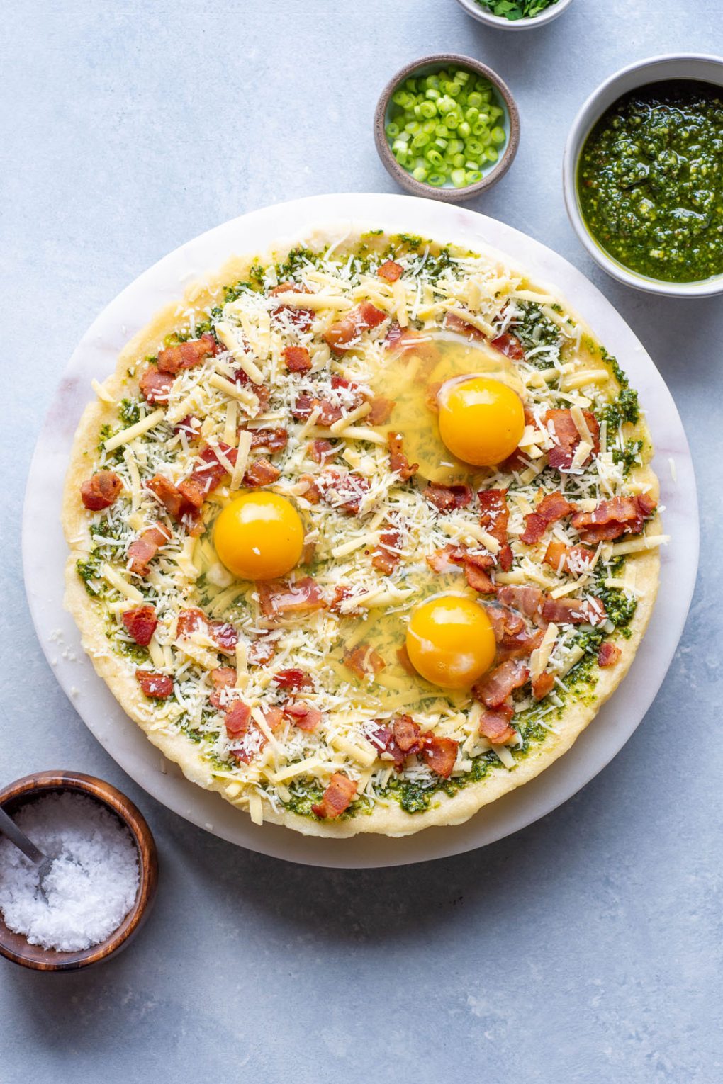 Overhead shot of an uncooked breakfast pizza. Topped with cheese, crispy bacon, raw sunny side up eggs, and fresh herbs and green onions. On a light colored marble serving tray and a light background. Next to a small bowl of sliced green onions, and another small bowl of pesto. 