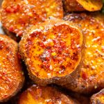 Close-up of roasted sweet potato rounds, seasoned with spices, chili flakes, and coarse salt, with a garnish of fresh cilantro visible in the corner. The potatoes have a golden-brown, crispy exterior.