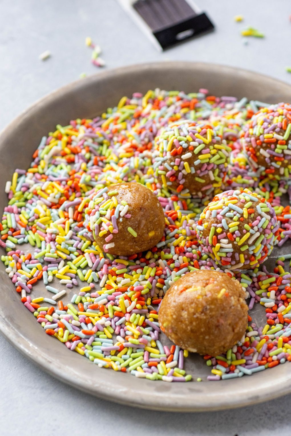 Side angle shot of energy bites on a small metal plate being rolled in rainbow sprinkles.