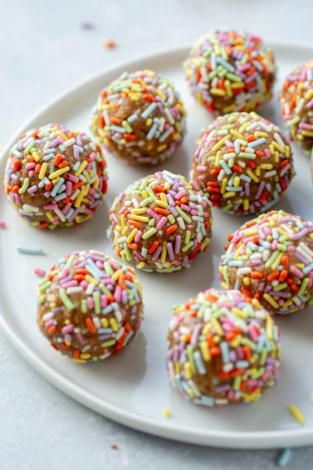 Close up side angle shot of energy bites coated in rainbow sprinkles on a white plate and white background surrounded by a few scattered sprinkles.