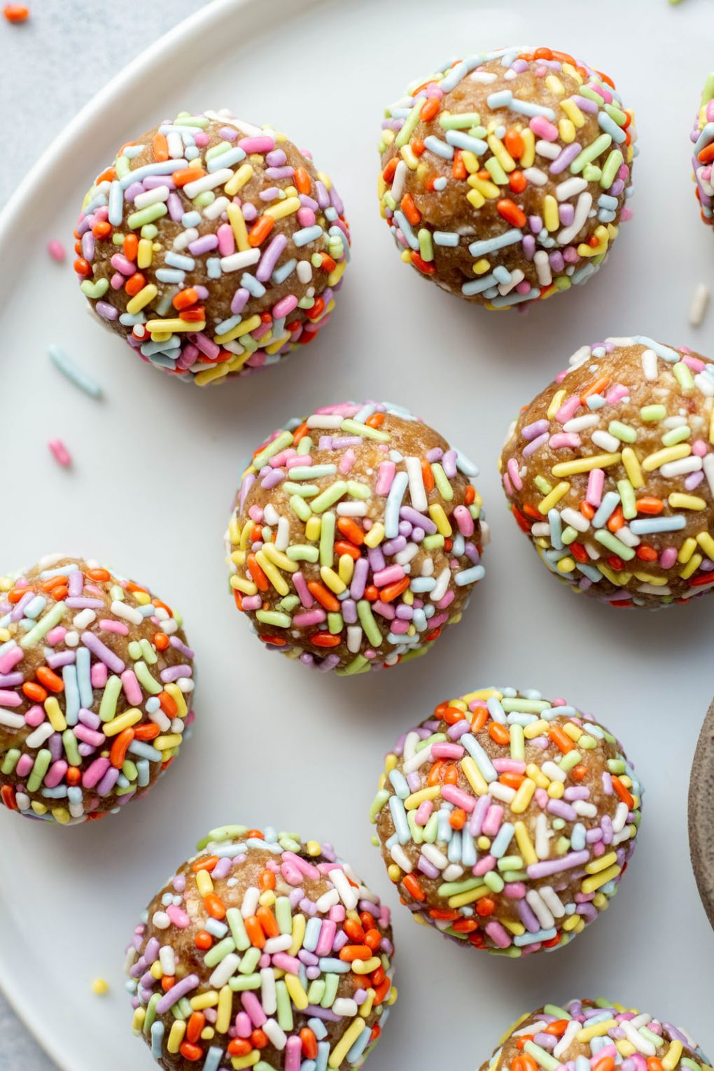 Overhead shot of energy bites coated in rainbow sprinkles on a white plate surrounded by a few scattered sprinkles.