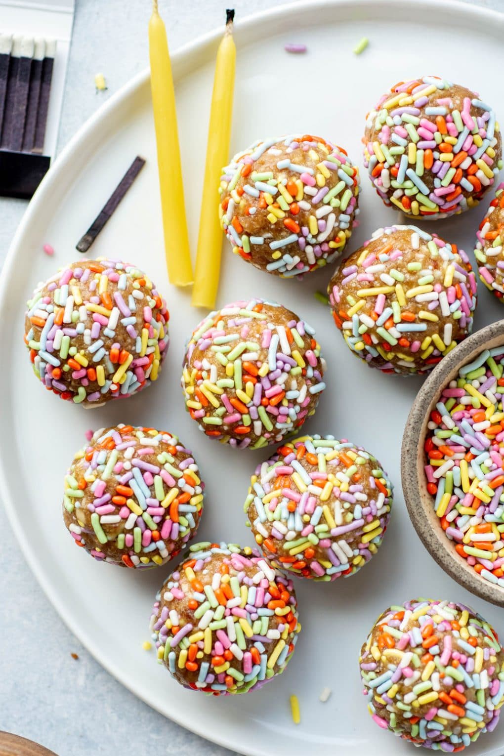 Overhead shot of energy bites coated in rainbow sprinkles on a white plate and white background, next to some beeswax birthday candles and scattered sprinkles.
