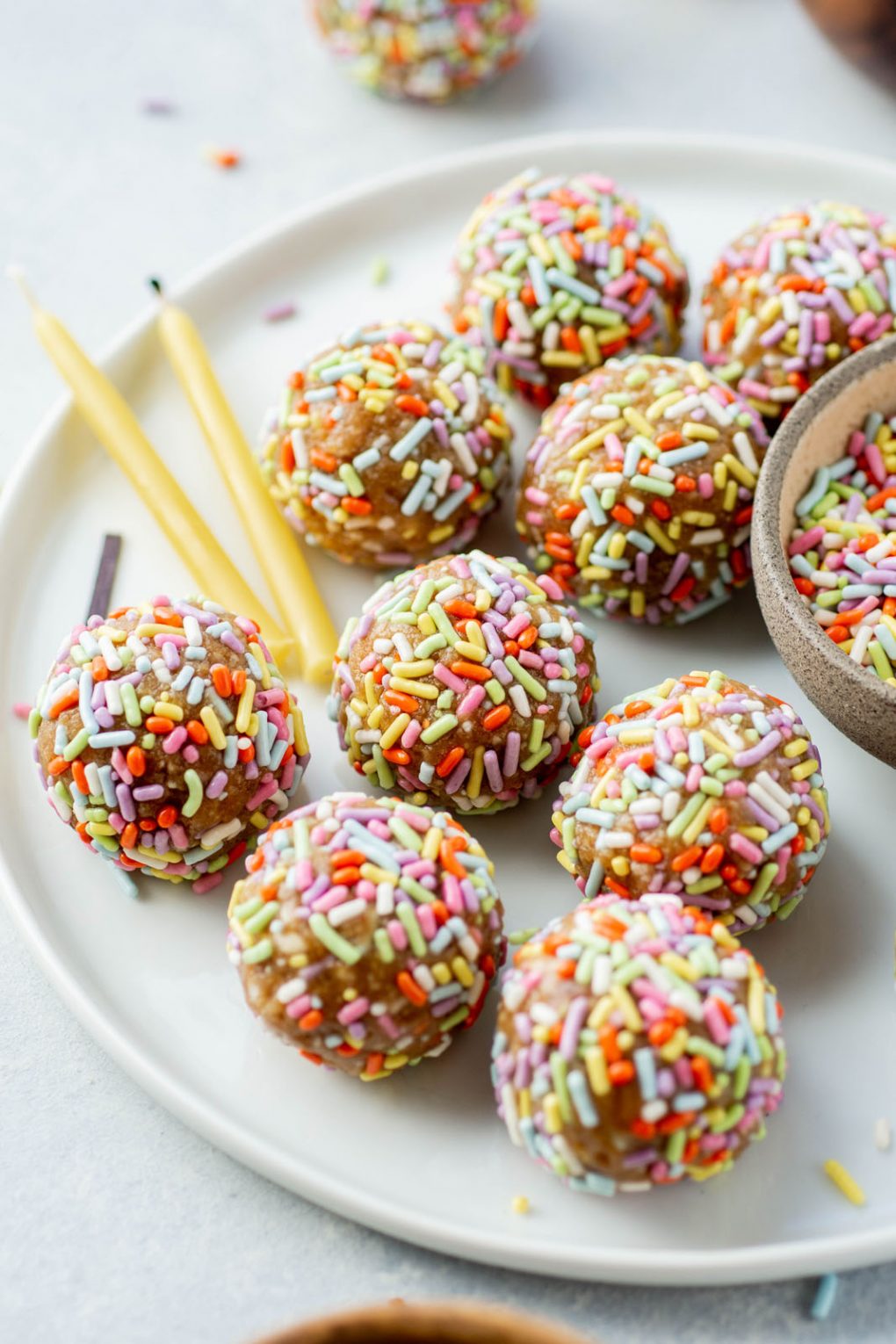 Side angle shot of energy bites coated in rainbow sprinkles on a white plate and white background, next to some beeswax birthday candles and scattered sprinkles.