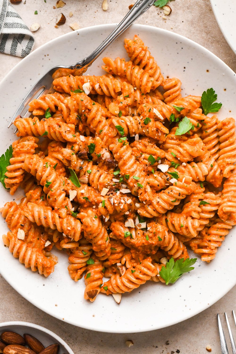 Romesco sauce tossed fusili pasta in a shallow speckled bowl, garnished with chopped roasted almonds and fresh parsley.
