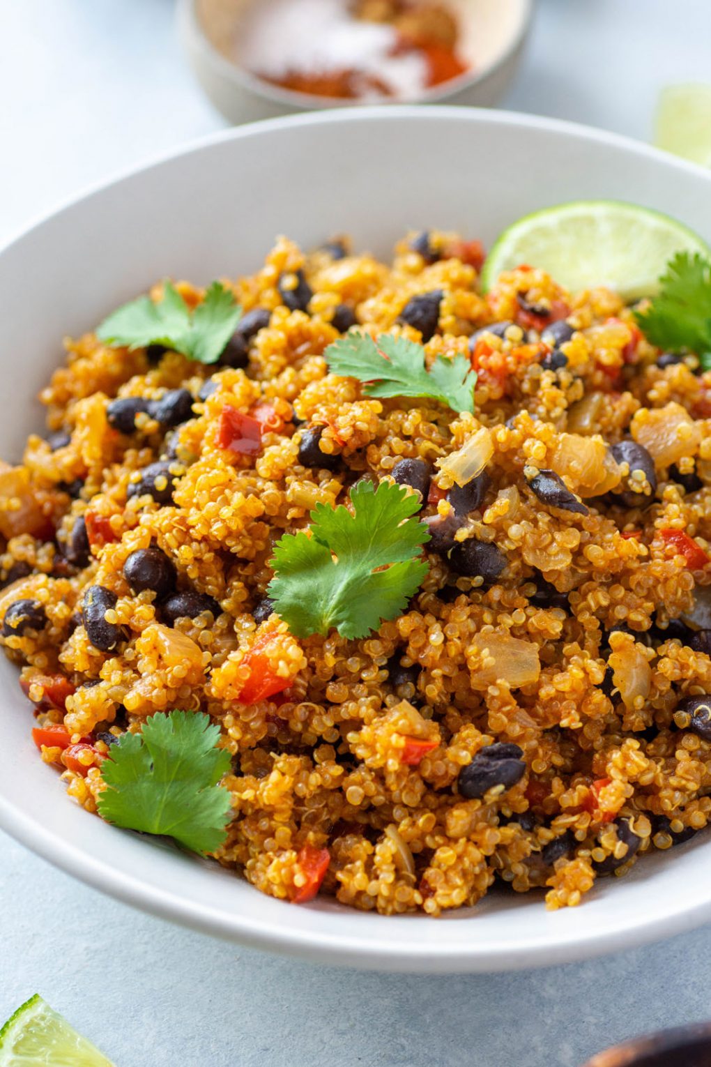 Close up side angle shot of southwest quinoa with black beans, diced red pepper, fresh cilantro, and garnished with lime wedges. In a white bowl on a light colored background. 